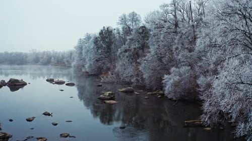 Flacher Fluss in der Nähe des Winterwaldes. Drohnenaufnahme verschneiter Bäume in der Nähe des Flusses.