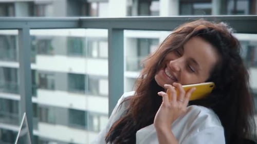 Happy Young Woman Talking on Mobile Phone While Sitting on Cozy Armchair on Home Terrace