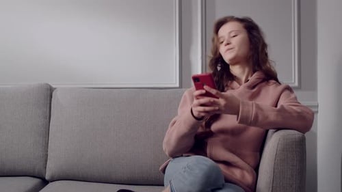 A Serious Girl Teenager Sits in a Room on a Sofa in a Cozy Home Environment and Uses a Smartphone A