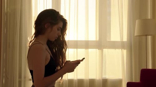 Happy Pretty Slim Young Woman in Black Top Sits and Scrolls on Phone in Hotel Room or at Home with