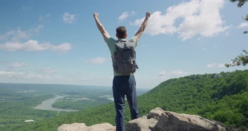 Man standing on top of a cliff looking at the view and raising his arms up in excitement for his ach