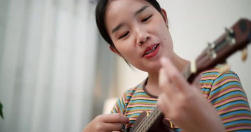 Beautiful young woman plays ukulele and sings in living room at home during holiday.