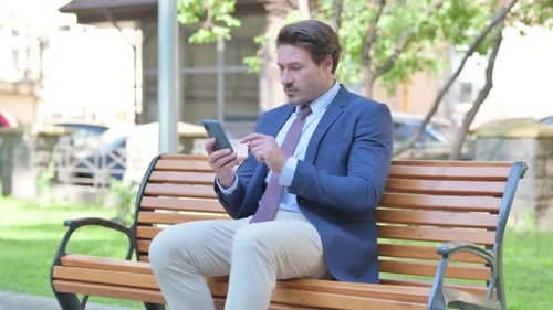 Man Using Smartphone on Park Bench in City