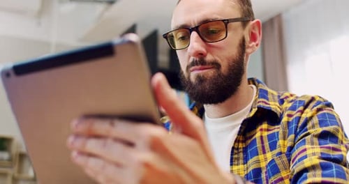 Close Up of Handsome Caucasian Young Man in Glasses Sitting on Couch and Using Tablet Device with