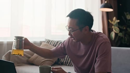 Man Pouring Tea in Cup while Speaking on Video by Laptop in Home Office