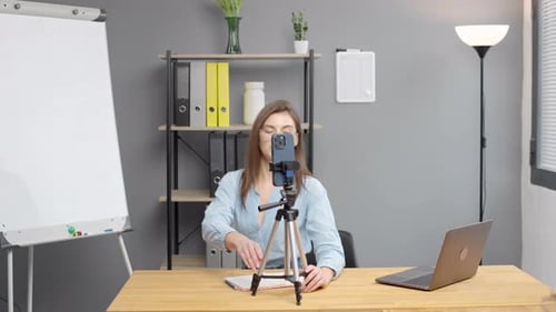 Smiling young female business blogger sitting at table in front of cellphone on stand