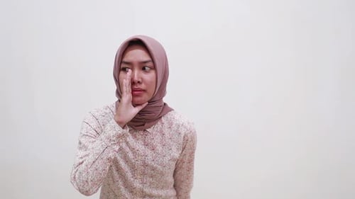 Young asian muslim woman standing over white background hand on mouth telling secret rumor, whisperi
