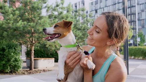 A Young Girl with a Happy Smile Stands Outdoors in a Park Area of the City Holds Pet a Jack Russell