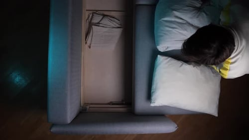 Un homme vu d'en haut dépliant un canapé-lit pour révéler le rangement avec des oreillers et de la literie préparant un