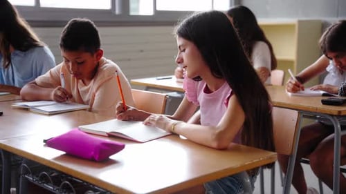 Diverse Primary School Children Concentrate on Writing in Notebooks While Sitting at Desk in Class
