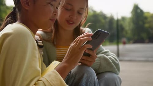 Close-up of Two Young Girls Talking and Looking at Smartphone Outdoors