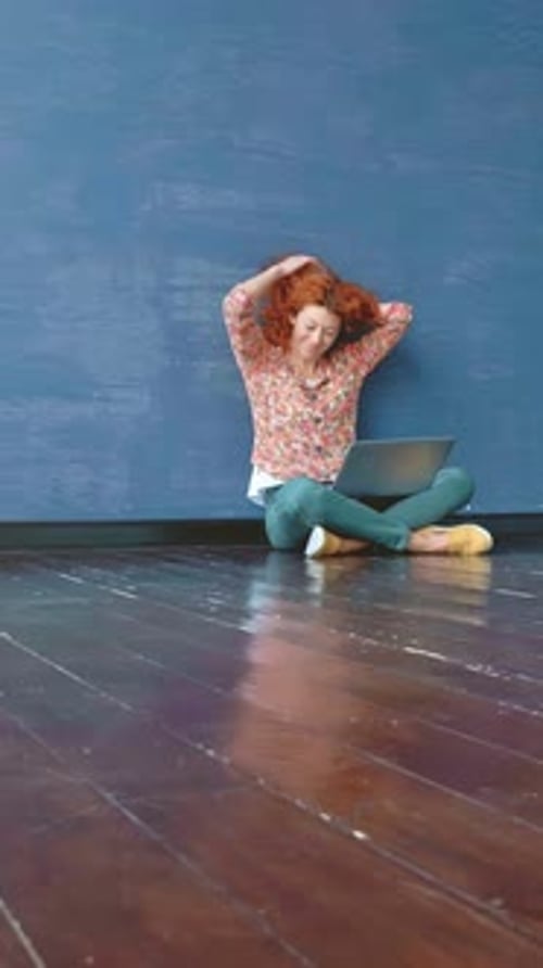 Woman with Curly Red Hair is Sitting on Wooden Floor Looking Frustrated While Working on Laptop