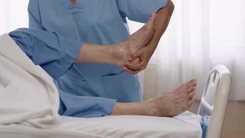 Asian nurse wearing blue scrubs helping elderly male patient with leg therapy on hospital bed