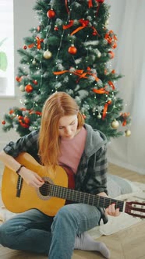 Young Woman Playing Guitar in Front of Christmas Tree Creating a Joyful Holiday Atmosphere at Home