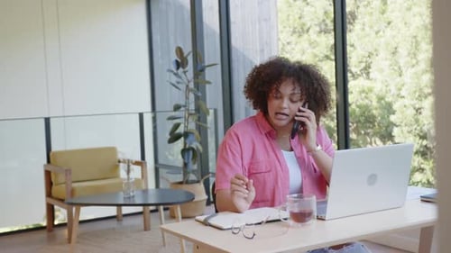 Talking on phone, woman working on laptop and writing in notepad at home office