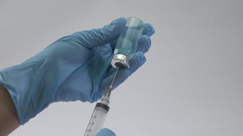 Medical gloves holding a syringe while drawing blue liquid from a glass vaccine vial