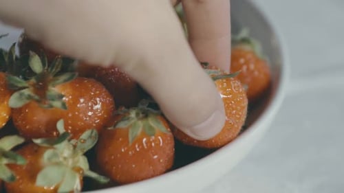 Detail of a white bowl of delicious red strawberries, a hand reaches in and takes one. Day light, sl