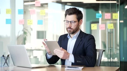 Businessman in a formal suit is using digital tablet sitting at the desk at workplace in business
