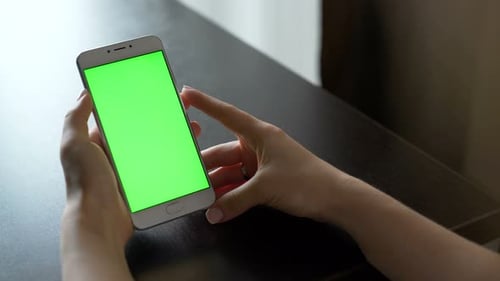 Young woman using touchscreen smartphone with green screen display indoors