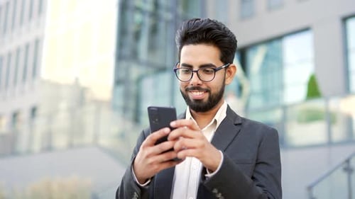 Smiling young businessman in formal suit is using a smartphone standing on street near an office
