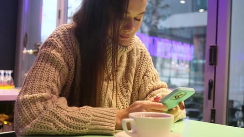 Young Pretty Woman in Sweater Sitting in Cafe with Coffee Using Phone Typing Chatting Window