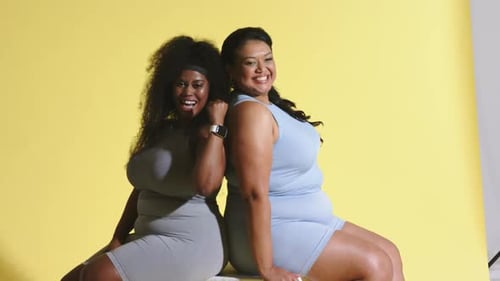 Diversas amigas posando espalda con espalda en un estudio amarillo flexionando los brazos para una sesión promocional