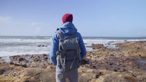 Young hiker with backpack and blue coat and red beanie knit hat is standing on rocky shore and looki