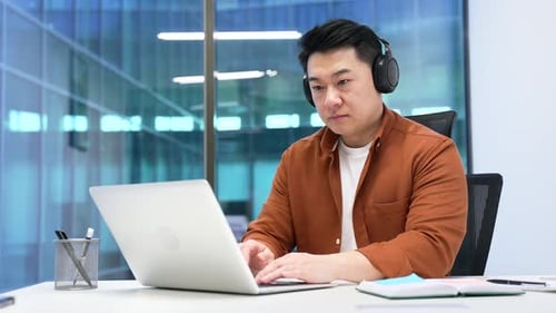 Confident Asian businessman in wireless headphones works on laptop sitting at workplace in business