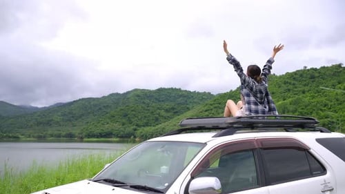 Asian woman sitting on car roof rack with raising arm up while travel on summer vacation road trip.