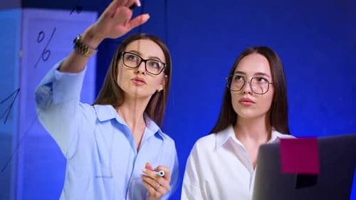 Two Caucasian brunette ladies wearing glasses stand at the glass board.