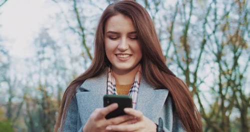 Close Up of Young Pretty Stylish Brunette Caucasian Woman Smiling and Chatting on Smartphone in Park