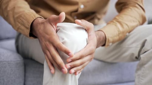 Close up Male hands massaging knee leg muscles while sitting on sofa in living room at home. Sad man