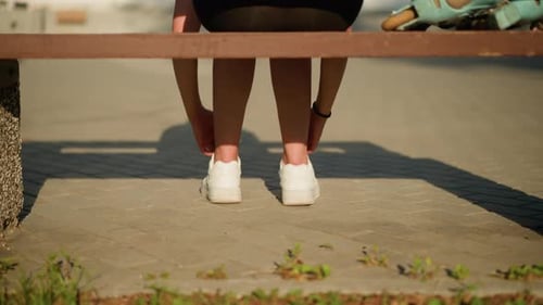 Lady Loosening Shoelaces of White Sneakers Seated on Bench in Bright Sunlight