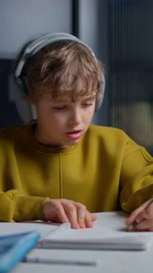 Young Boy Wearing Large Headphones Intensely Focused on Studying and Writing in Notebooks While