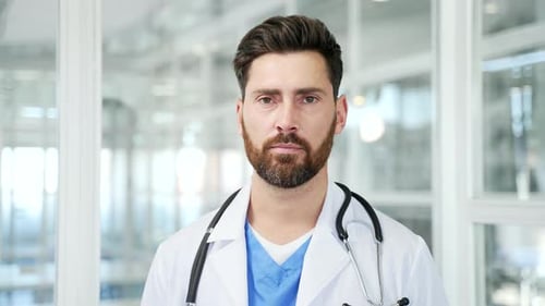 Portrait of serious doctor in white coat standing in modern hospital clinic looking at camera.