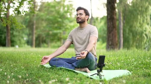 Young happy sportsman practices yoga on mat in urban city park using an application on his mobile