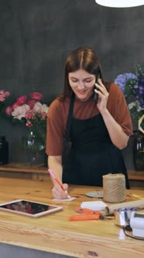 Young Beautiful Woman Florist in an Apron in a Flower Shop Receives an Order By Phone to Make a