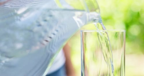 A Woman Pouring Pure Water Into Glass From Jug on Green Nature Background