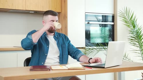 joven apuesto barbudo trabajando por la mañana en casa en una mesa con ordenador portátil en la cocina