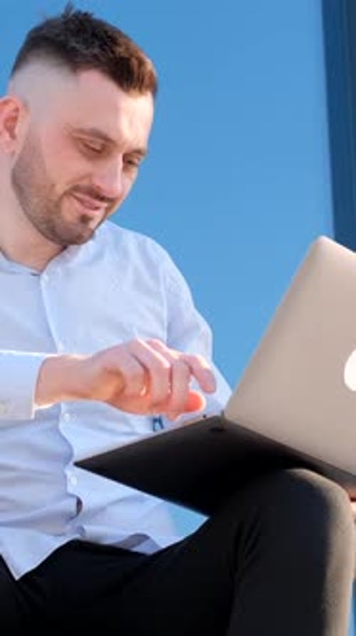 A Handsome Man is Sitting Outside Near an Office Center and Working on a Laptop Vertical Video