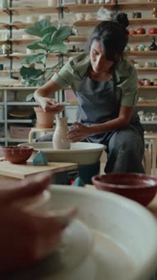 Hispanic Woman Making Clay Vase on Potters Wheel in Workshop