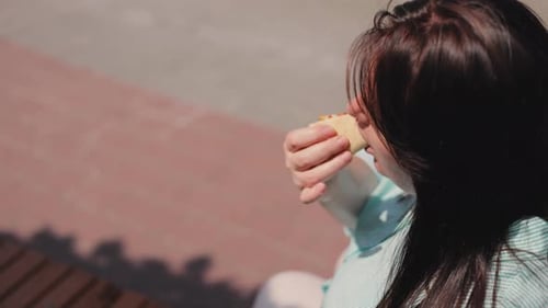 A brown-haired girl in a striped shirt sits on the wooden bench in the park and eats pizza