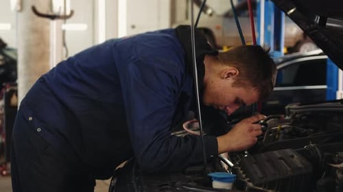 Young auto mechanic under the hood of a car with a flashlight checks the condition of the engine