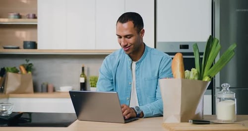 Man Uses Laptop in Bright Kitchen With Groceries