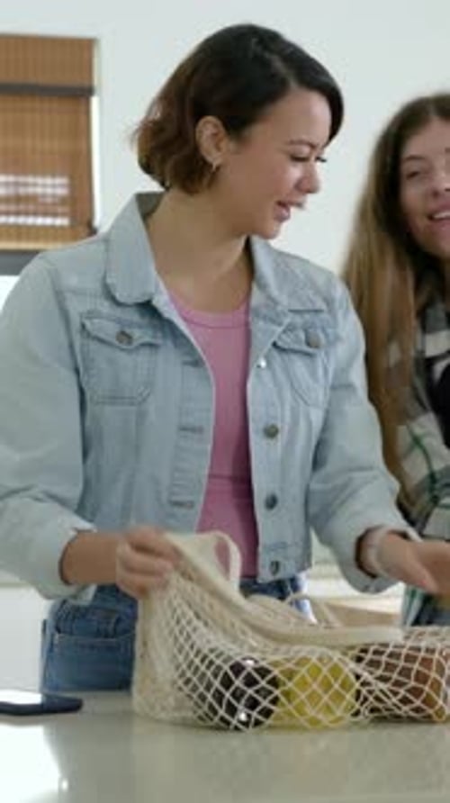 Vídeo vertical: metiendo la mano en una bolsa de malla, una mujer vestida de mezclilla desempaca la comida en la cocina y coloca una botella