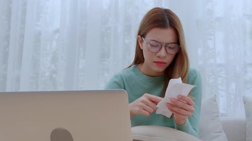Young asian woman wearing glasses sitting on sofa worried with bills about financial and stress.