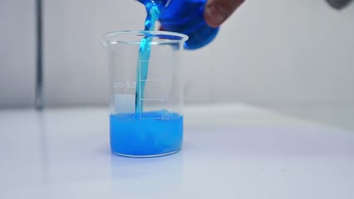 Hand of researcher pours blue liquid into the beaker with diaper filler. Blue liquid mixes up