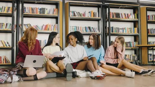 Five Happy Multiethnic Female Students Sit on the Floor in a University Library Chatting and