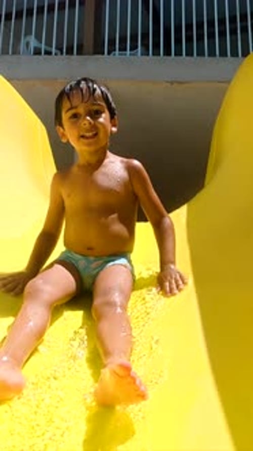 Happy Child Sliding Into Swimming Pool on Yellow Slide