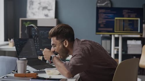 Young Indian Male Employee Eating Lunch during Coding on Laptop in Office